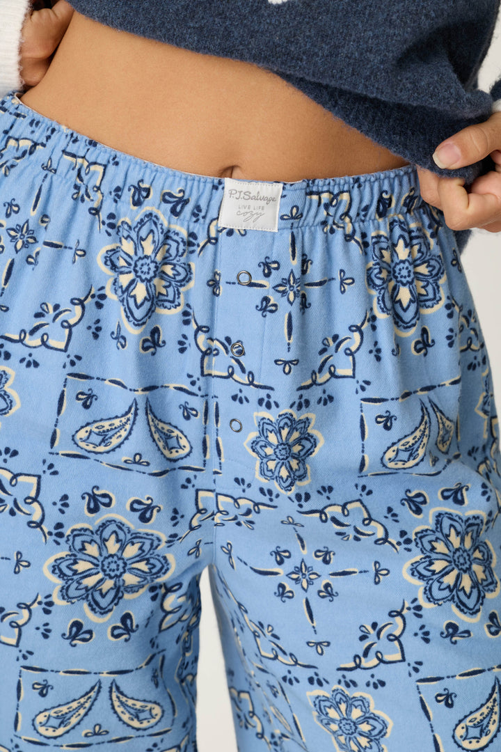 A person is wearing the Classic Paisley Bandana Print Flannel Pajama Pants with a blue floral and paisley design, holding the waistband to display the P.J. Salvage brand tag.