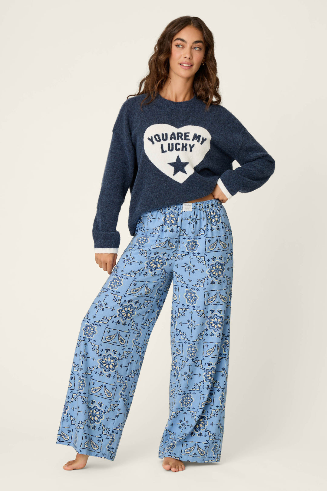 A person stands barefoot in Classic Paisley Bandana Print Flannel Pajama Pants by P.J. Salvage and a navy sweater with a white heart that reads “YOU ARE MY LUCKY” and features a star. They have one hand on their hip and are smiling slightly.