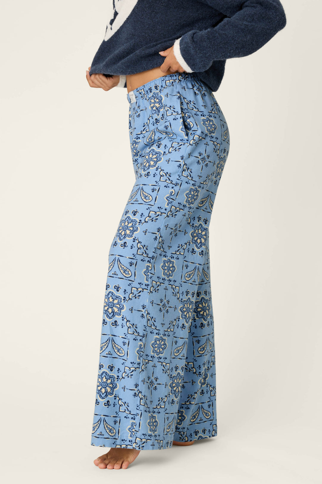 A person is seen barefoot and standing sideways against a plain background, wearing the P.J. Salvage Classic Paisley Bandana Print Flannel Pajama Pants in light blue with a dark blue floral and paisley design, paired with a navy sweater.