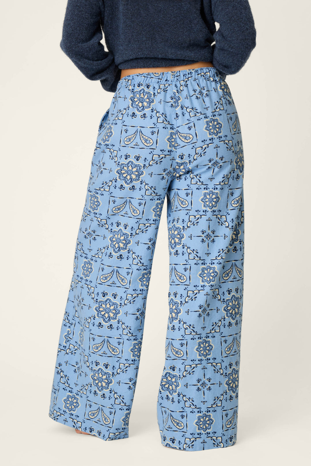 Person wearing P.J. Salvage Classic Paisley Bandana Print Flannel Pajama Pants, featuring a loose fit and light blue fabric with dark blue paisley and geometric designs, standing with hands on hips and photographed from the back against a plain background.