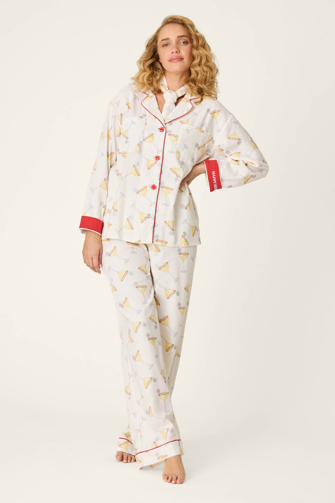 A woman stands and smiles, wearing the P.J. Salvage Happy Hour Cocktail Print 3-Piece Flannel PJ Set. The white pajama top features red trim, red buttons, and red cuffs, while the loose-fitting pants complete the set—all adorned with a playful cocktail print. The background is plain white.
