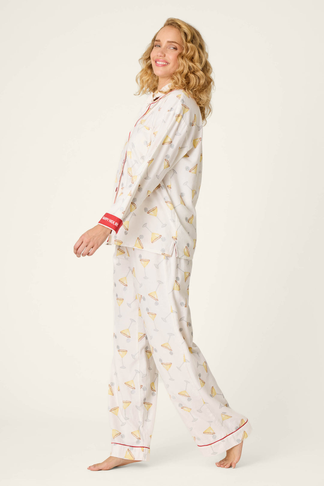 A woman with curly blonde hair smiles while wearing the P.J. Salvage Happy Hour Cocktail Print 3-Piece Flannel PJ Set, featuring a playful martini glass pattern and red trim. She stands barefoot and sideways against a plain light background.