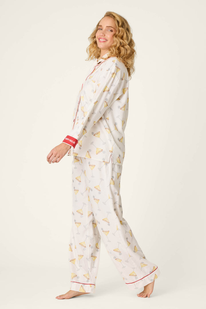 A woman with curly blonde hair smiles while wearing the P.J. Salvage Happy Hour Cocktail Print 3-Piece Flannel PJ Set, featuring a playful martini glass pattern and red trim. She stands barefoot and sideways against a plain light background.