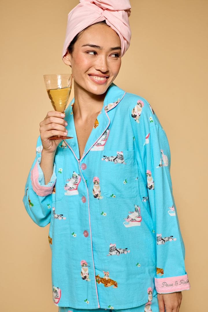 A woman wearing a light blue pajama set with a pink towel wrap in her hair and holding a glass of champagne, set against a solid beige background.