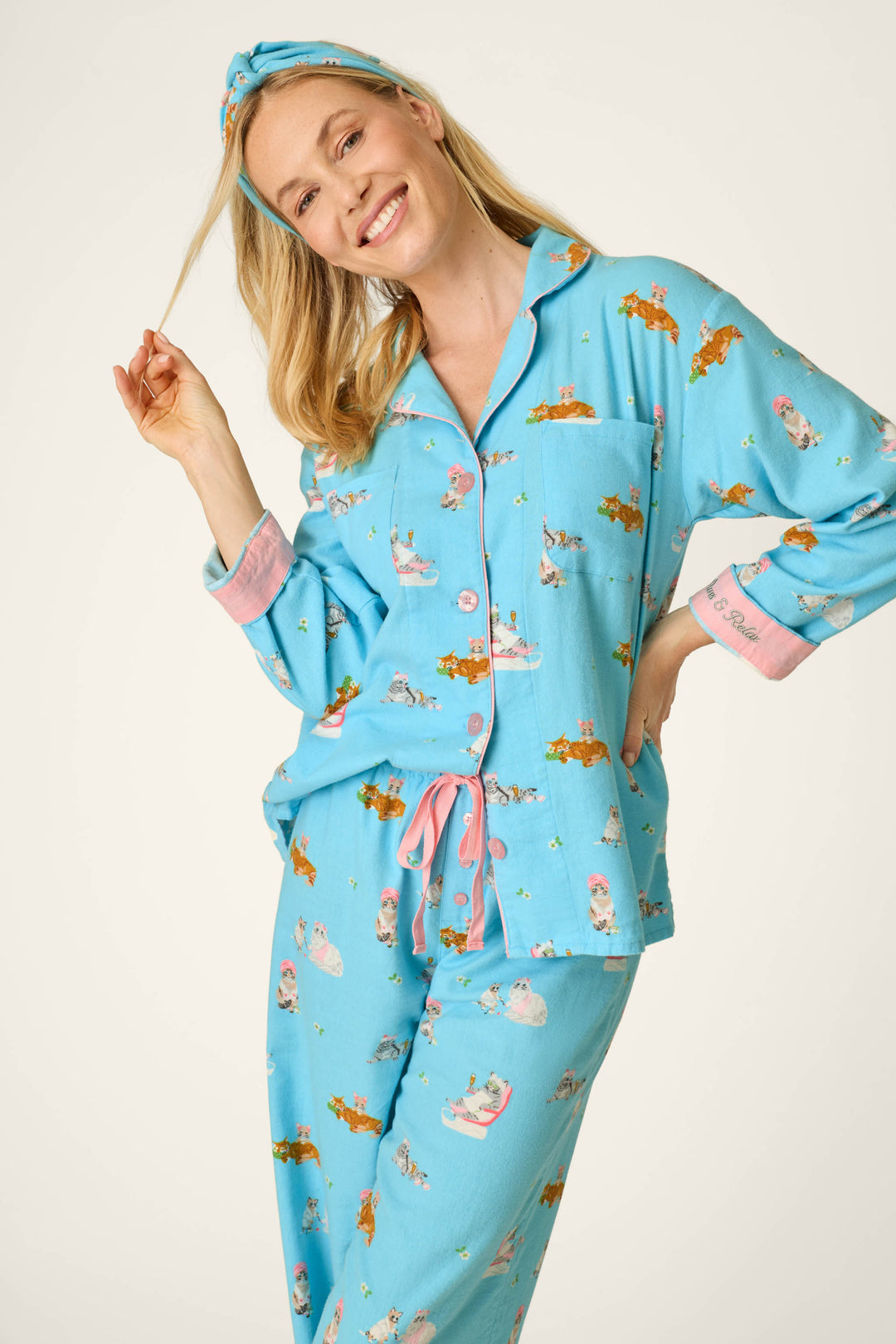 A smiling woman wearing the P.J. Salvage Pampered Cat Print 3-Piece Long Sleeve Flannel PJ Set and a matching headband holds a strand of her blonde hair, standing against a light, plain background.