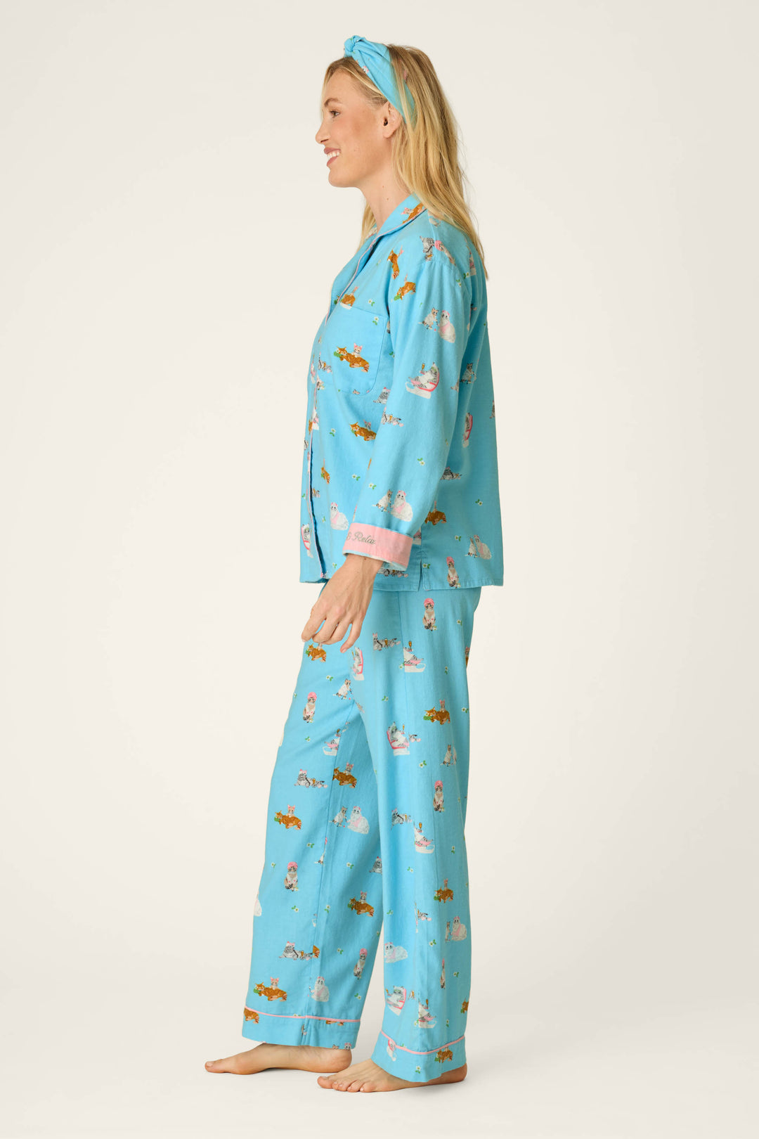 A woman in profile stands barefoot against a plain background, wearing the P.J. Salvage Pampered Cat Print 3-Piece Long Sleeve Flannel PJ Set with colorful cat patterns and a matching blue headband.
