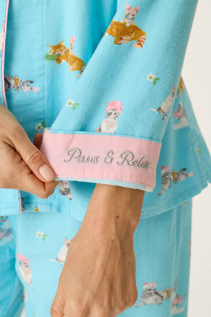 Wearing the Pampered Cat Print 3-Piece Long Sleeve Flannel PJ Set from P.J. Salvage, a person holds a sleeve featuring pink trim embroidered with the words “Paws & Relax.”.