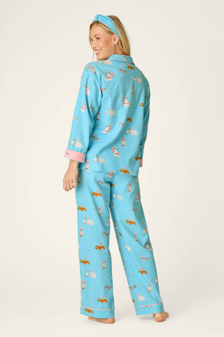 A person stands barefoot, smiling and glancing over their shoulder. They are wearing the P.J. Salvage Pampered Cat Print 3-Piece Long Sleeve Flannel PJ Set, featuring playful cat prints and a coordinating blue headband, set against a plain light background.