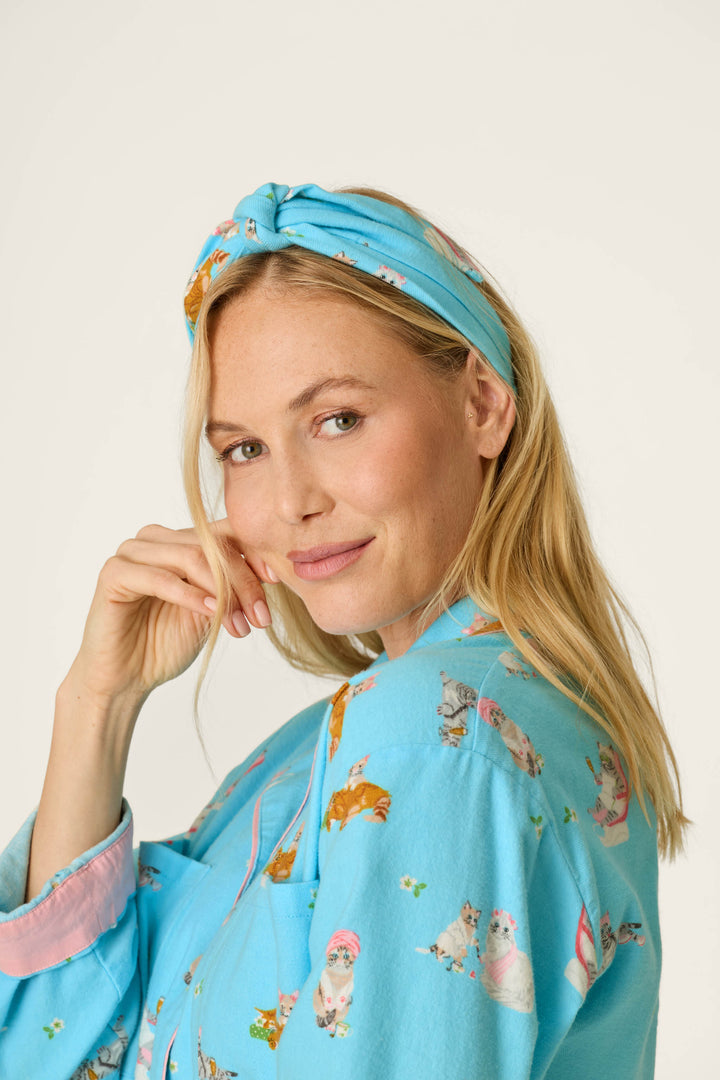 A woman with blonde hair smiles gently at the camera against a plain background, wearing the P.J. Salvage Pampered Cat Print 3-Piece Long Sleeve Flannel PJ Set—blue pajamas adorned with colorful cat illustrations and a coordinating blue headband.