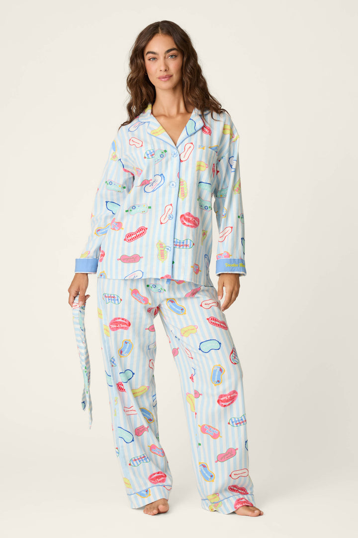 A woman wears the P.J. Salvage Out of Office Eyemask Print 3-Piece Flannel PJ Set, featuring a light blue, loose-fitting pajama set decorated with colorful lips, toothbrushes, toothpaste, and mouth prints. She stands against a plain background holding the matching fabric belt.