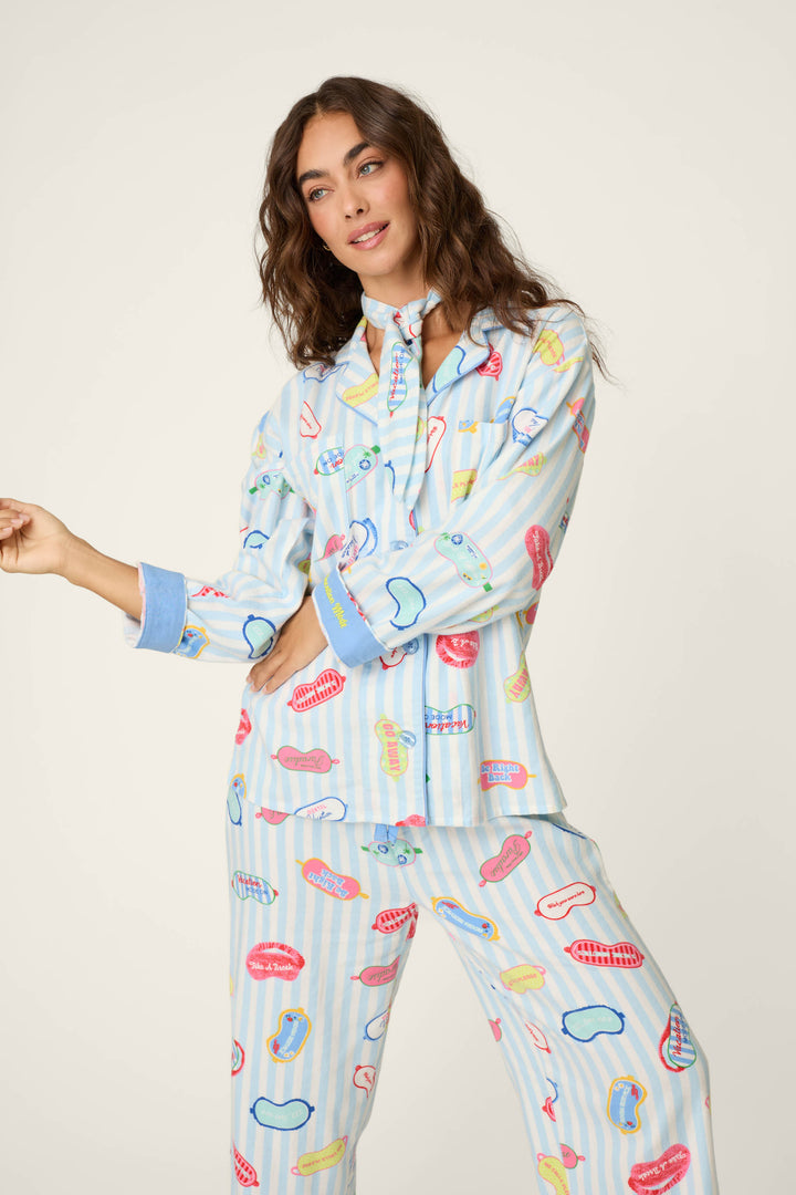 A woman stands against a plain background with her head slightly tilted, wearing the P.J. Salvage Out of Office Eyemask Print 3-Piece Flannel PJ Set in a light blue striped pattern decorated with colorful cartoon mouths and lips.