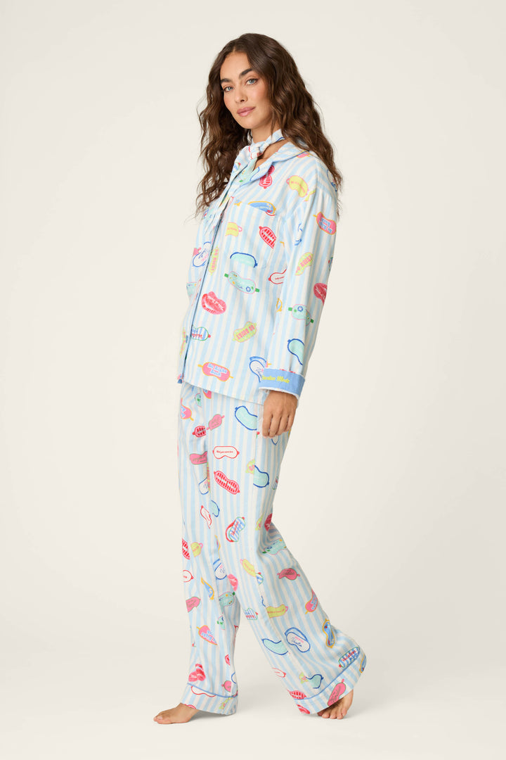 A woman with long wavy hair models the P.J. Salvage Out of Office Eyemask Print 3-Piece Flannel PJ Set—loose, light blue pajamas adorned with colorful illustrations of lips, toothbrushes, and toothpaste. She stands barefoot against a plain light background, looking toward the camera.