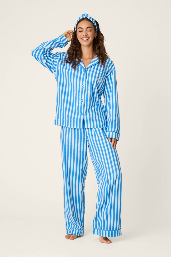 A woman smiles with her eyes closed, wearing the P.J. Salvage Snowflake Icon 3-Piece Long Sleeve Flannel PJ Set, featuring blue and white stripes and a matching headband, as she stands barefoot against a plain light background.