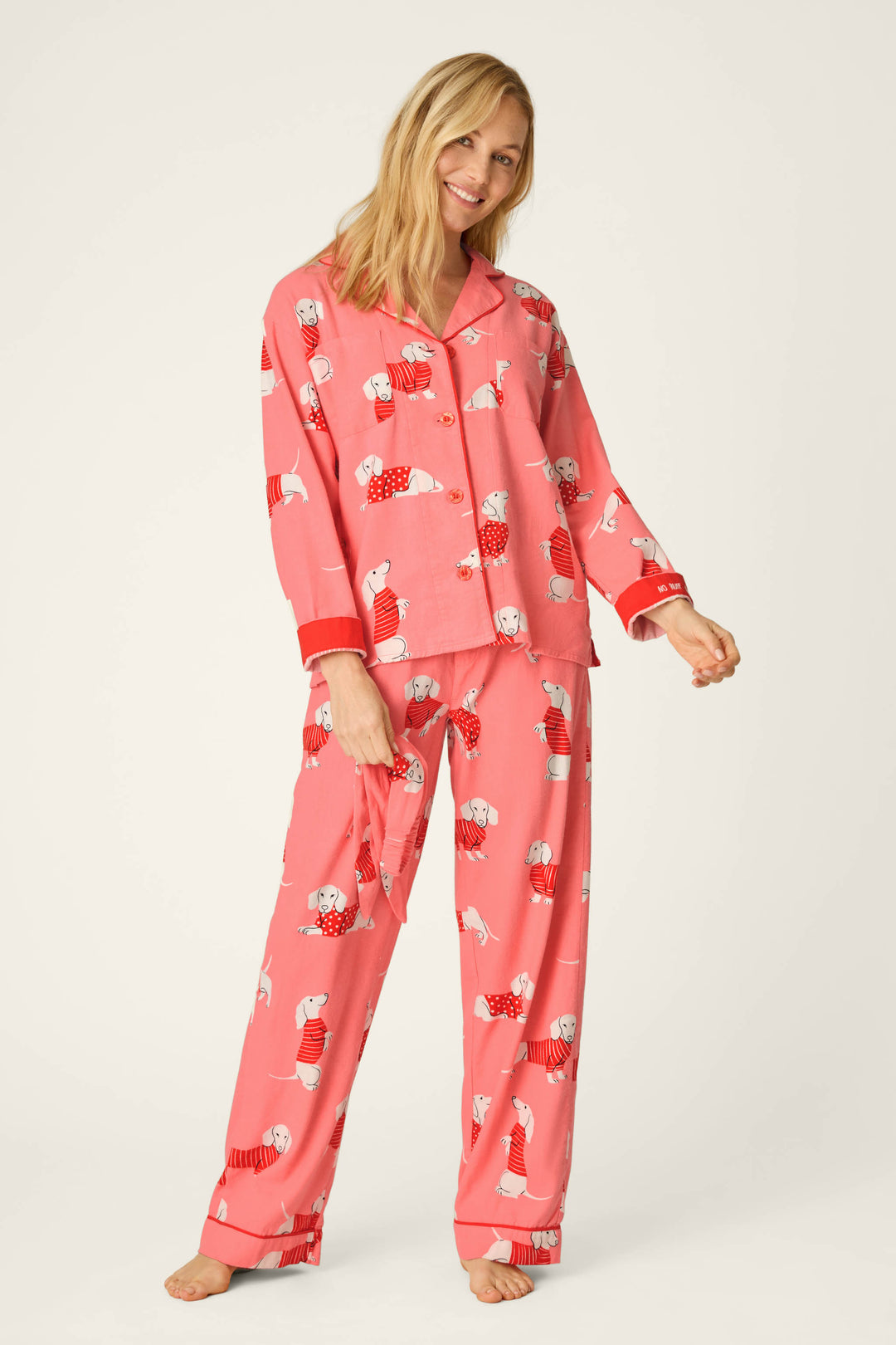 A woman stands barefoot and smiling against a plain background, wearing the P.J. Salvage Dachshund Print 3-Piece Flannel PJ Set. The set features pink pajamas adorned with playful dachshund dog illustrations and red cuffs, including a button-up top and loose pants.