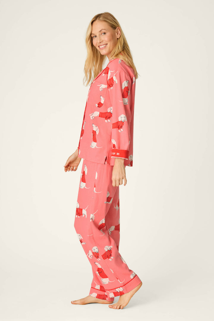 A woman stands barefoot, smiling, and wearing the P.J. Salvage Dachshund Print 3-Piece Flannel PJ Set featuring a playful dachshund pattern with red accents. She is posing sideways against a plain light background.