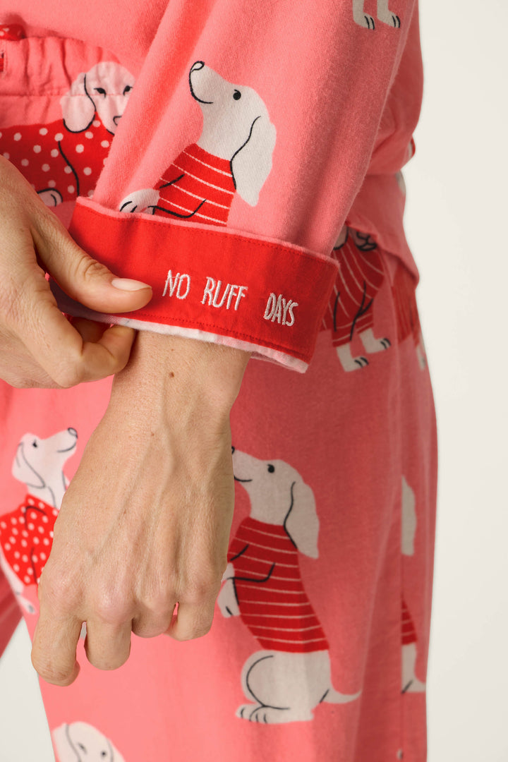 Wearing the P.J. Salvage Dachshund Print 3-Piece Flannel PJ Set, a person holds the sleeve, which is printed with “NO RUFF DAYS.” The pink pajamas feature a playful pattern of white dachshunds in red sweaters.
