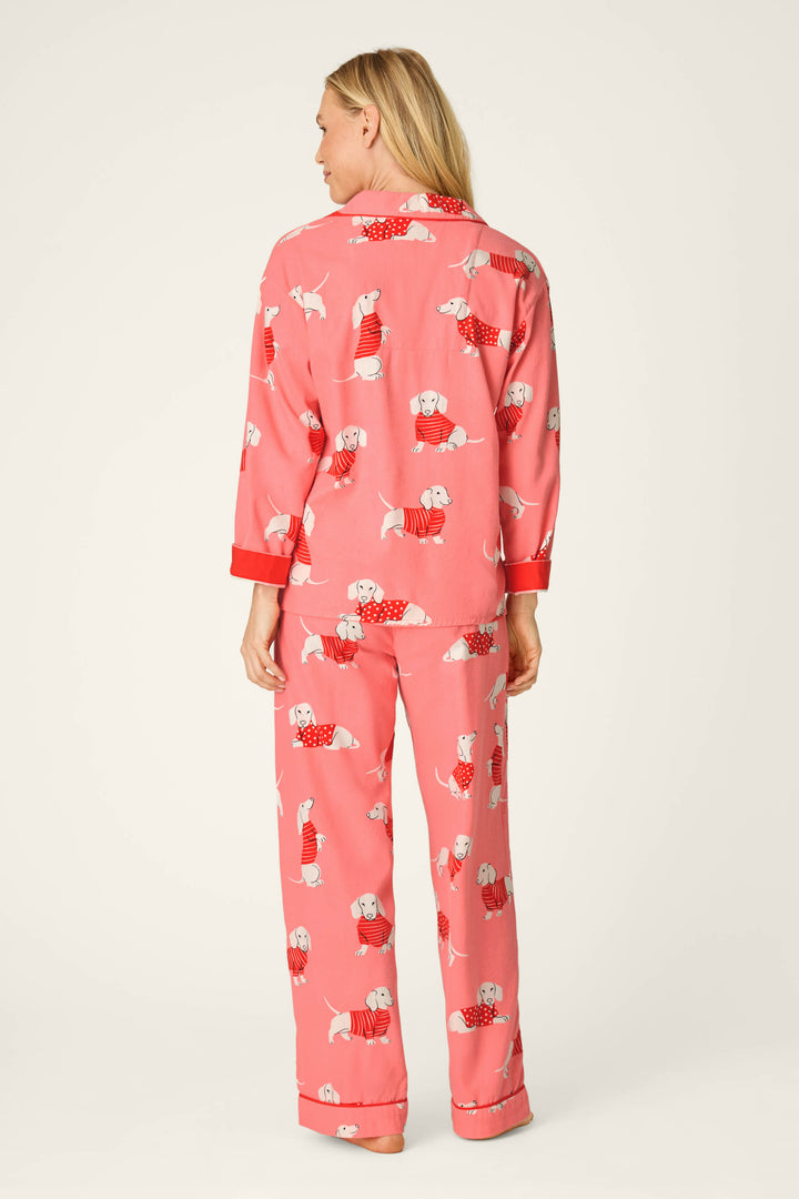 A woman stands barefoot, facing away, wearing the P.J. Salvage Dachshund Print 3-Piece Flannel PJ Set. The pajamas feature a playful dachshund print in pink, with a long-sleeve top and loose-fitting pants. The background is plain and light-colored.