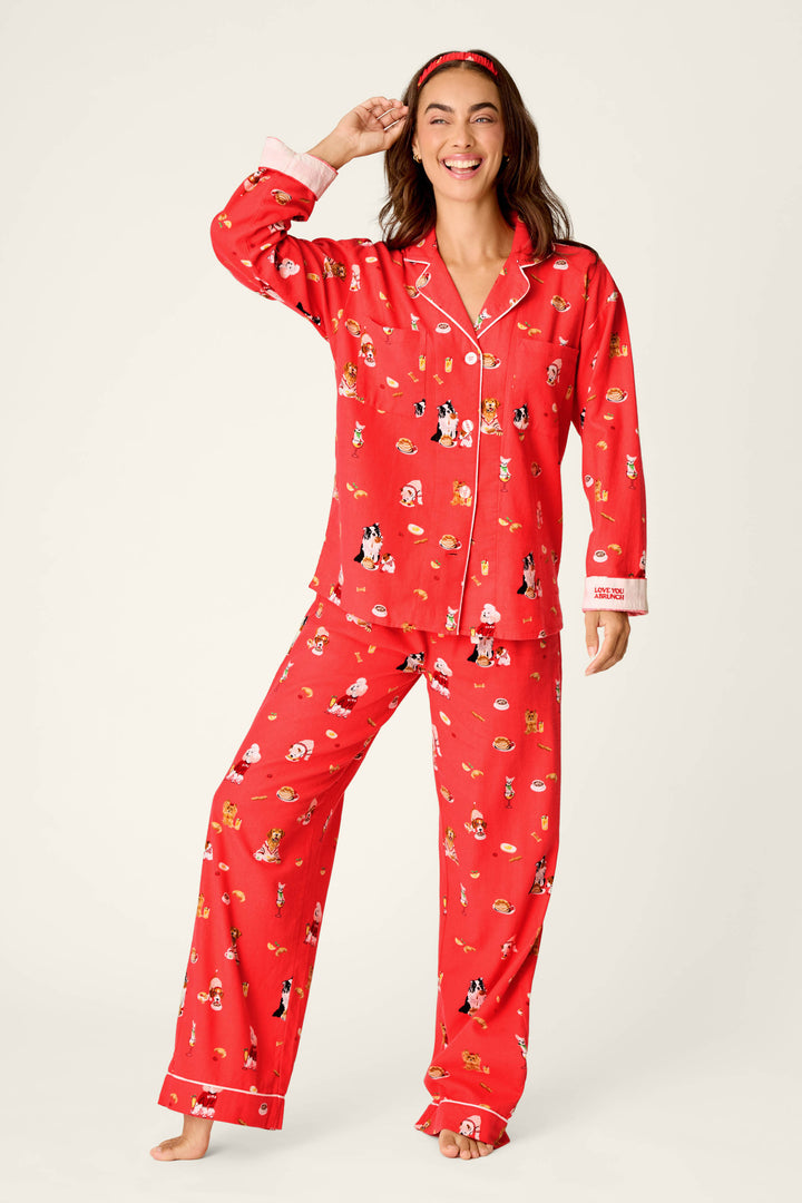 A woman wearing the P.J. Salvage “Dogs Who Brunch” Print 3-Piece Long Sleeve Flannel PJ Set, featuring playful dog cartoon prints and white piping with text detail on the sleeve, stands barefoot, smiling and touching her head, complemented by a matching red headband.