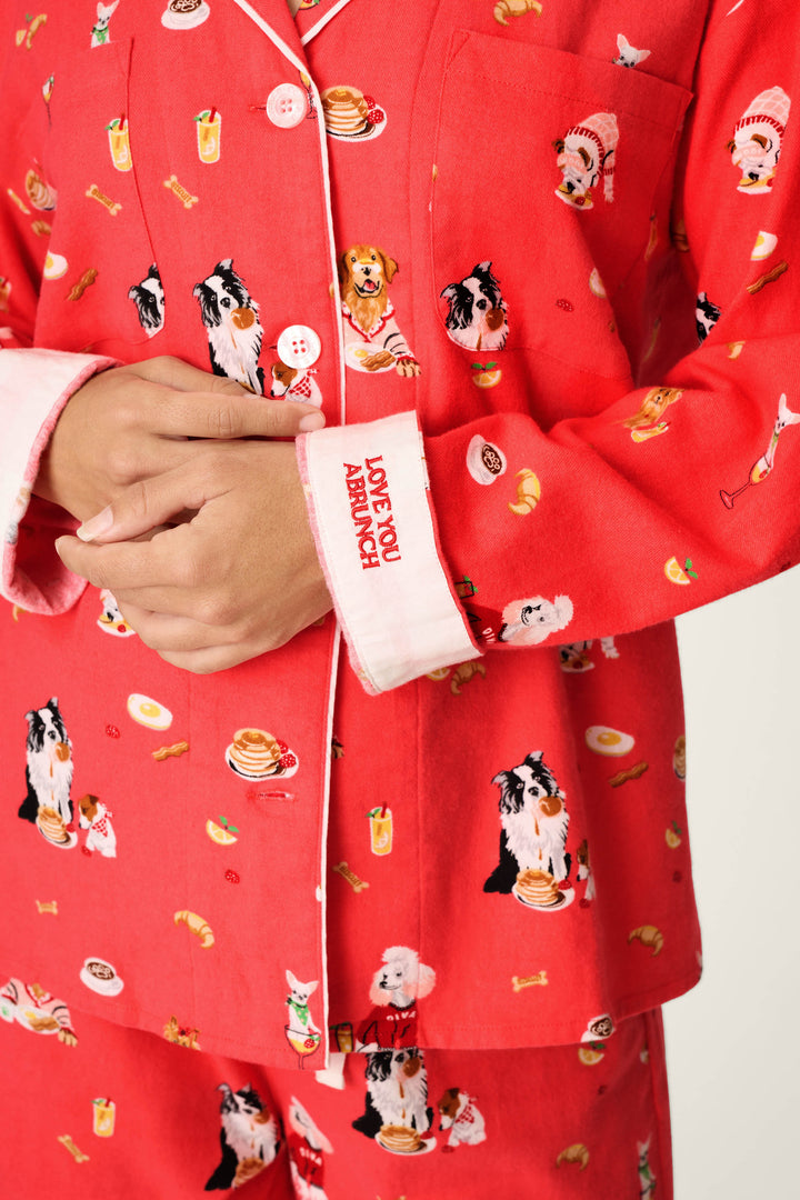 A person wearing the P.J. Salvage Dogs Who Brunch Print 3-Piece Long Sleeve Flannel PJ Set, featuring red pajamas adorned with dogs and breakfast foods, has their hands folded. The sleeve cuff displays “LOVE YOU A BRUNCH” in red text.
