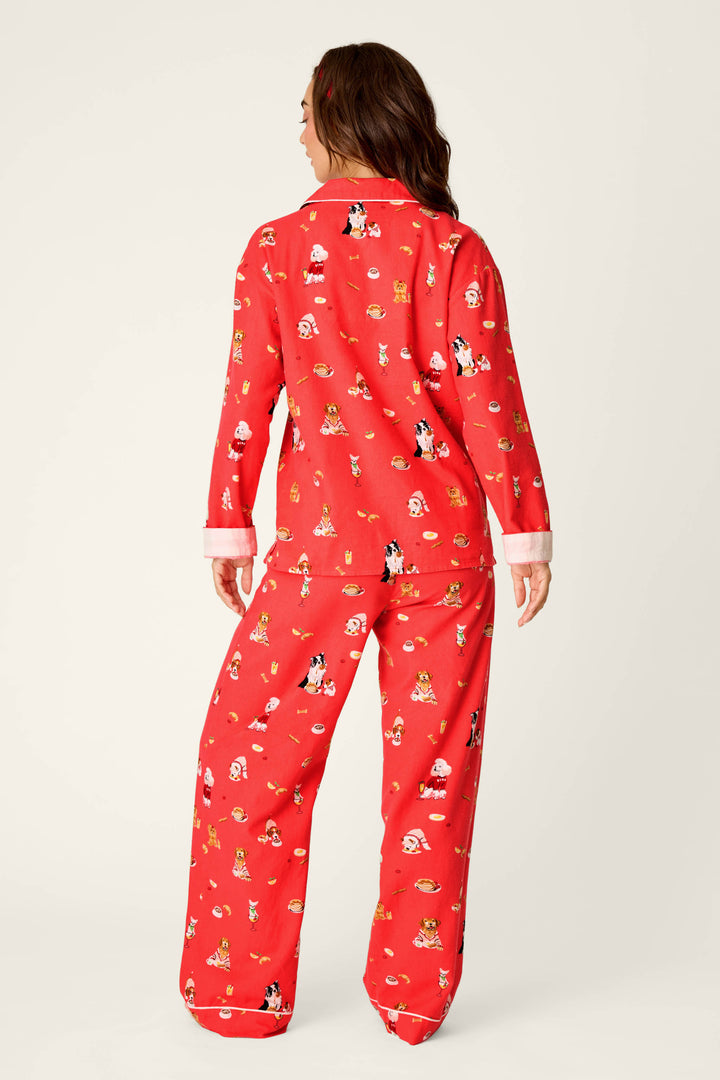 A woman stands facing away, wearing the P.J. Salvage Dogs Who Brunch Print 3-Piece Long Sleeve Flannel PJ Set. The red pajamas feature playful cartoon dog prints, white cuffs, and a relaxed fit. The background is plain and light-colored.