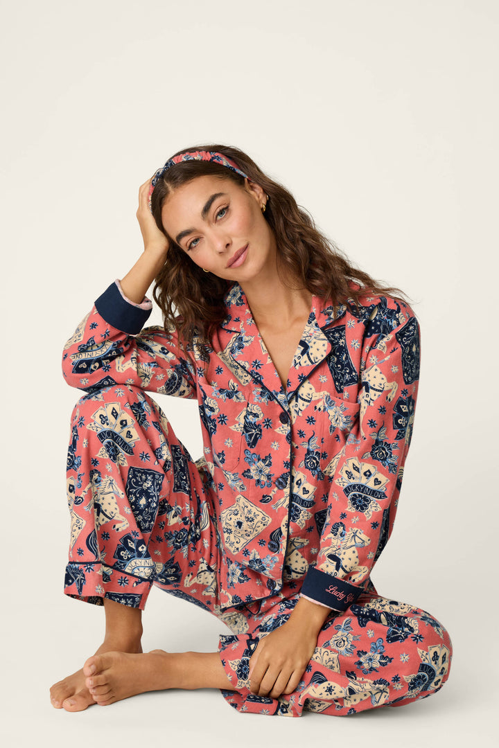 A woman with long brown hair sits barefoot in the P.J. Salvage Lucky in Love Western Print 3-Piece Long Sleeve Flannel PJ Set, including a matching headband, resting her head on her hand against a plain light background.