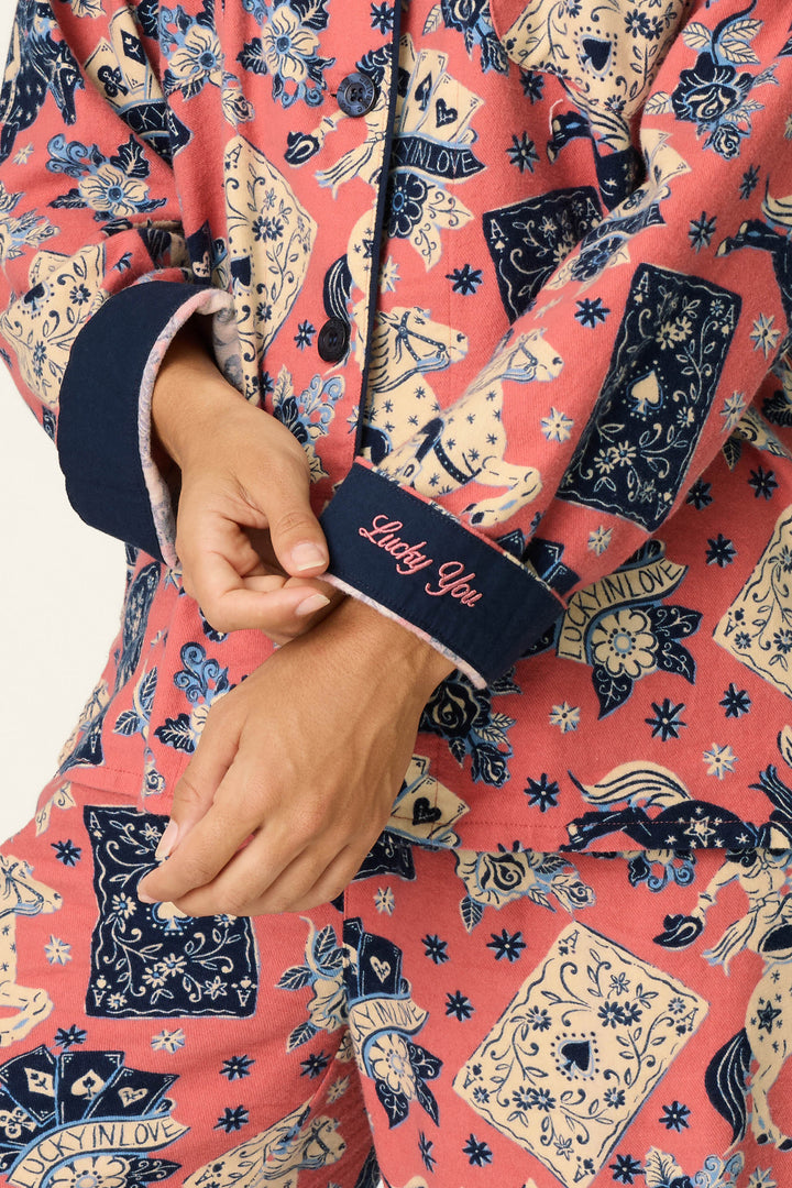 Wearing the P.J. Salvage Lucky in Love Western Print 3-Piece Long Sleeve Flannel PJ Set, a person displays the navy cuff embroidered with “Lucky You” in pink script. The colorful pajamas feature western-inspired card and floral patterns, detailed with hearts, stars, and the phrase “LUCKY IN LOVE.”.