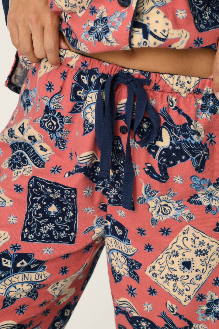 Close-up of a person wearing the P.J. Salvage Lucky in Love Western Print 3-Piece Long Sleeve Flannel PJ Set, featuring pink pajama pants with blue and white tarot card and skull patterns, a blue drawstring at the waist, and the person’s hands resting at the waistband.