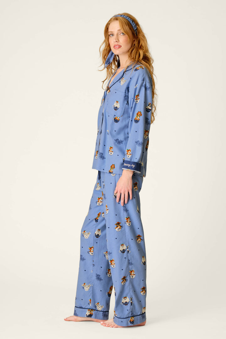 A woman with long hair stands barefoot and slightly to the side against a plain background, wearing the P.J. Salvage “Teacup Pups” 3-Piece Flannel PJ Set—a matching blue pajama set featuring a playful cartoon dog print and a coordinating headband.