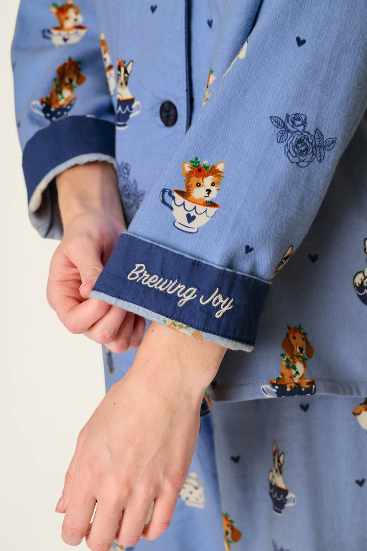 Close-up of a person wearing the P.J. Salvage Teacup Pups 3-Piece Flannel PJ Set, featuring blue pajamas printed with adorable dogs in teacups and a cuff embroidered with “Brewing Joy” in white script.
