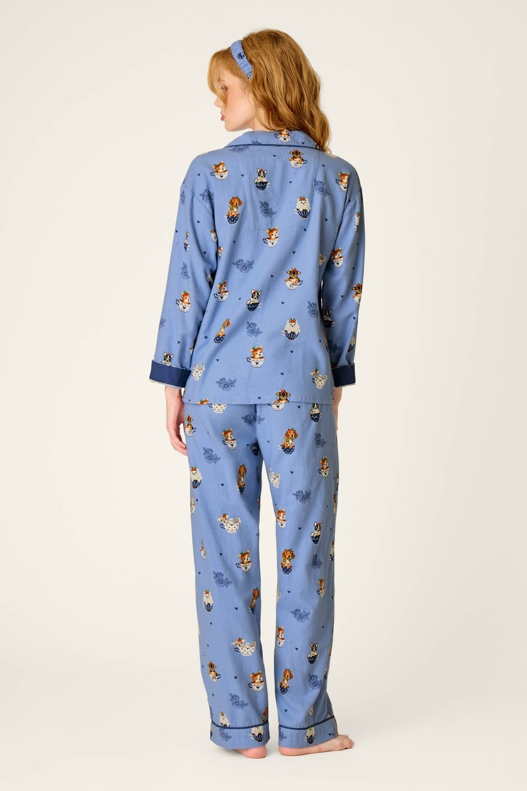 A person with long hair is seen from behind wearing the P.J. Salvage Teacup Pups 3-Piece Flannel PJ Set—blue pajamas featuring a cartoon dog print—with bare feet and a matching blue headband, against a simple light background.