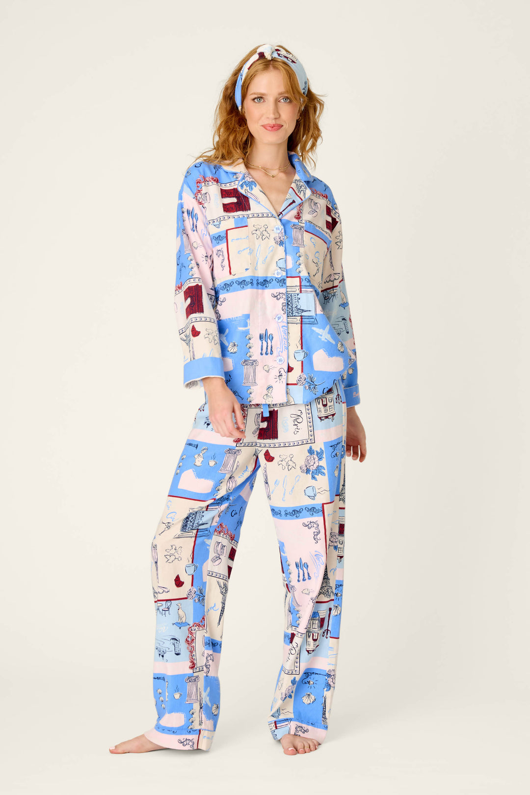 A woman stands barefoot against a plain light background, wearing the P.J. Salvage Allons-y Parisian Print 3-Piece Flannel PJ Set featuring a whimsical dog and kitchen motif in coordinating blue and cream tones, complete with a matching headband.