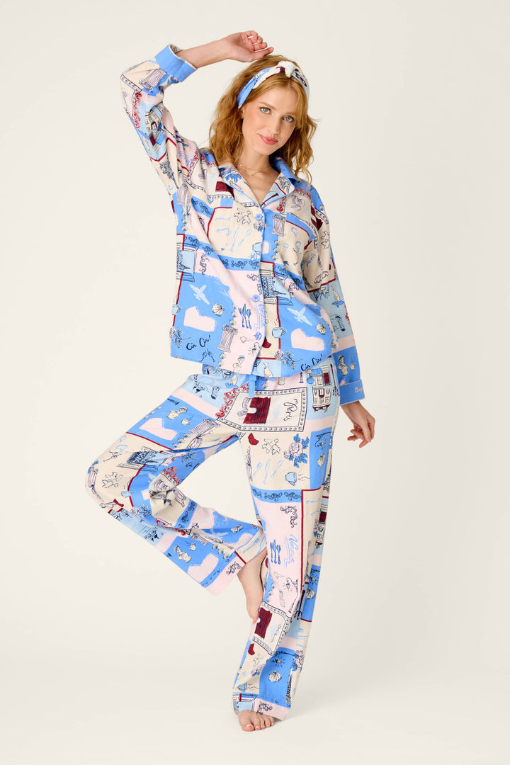 A woman models the P.J. Salvage Allons-y Parisian Print 3-Piece Flannel PJ Set, featuring vibrant blue and cream illustrated patterns. She poses barefoot on one leg with one arm raised and her other hand resting on her head against a simple, light backdrop.