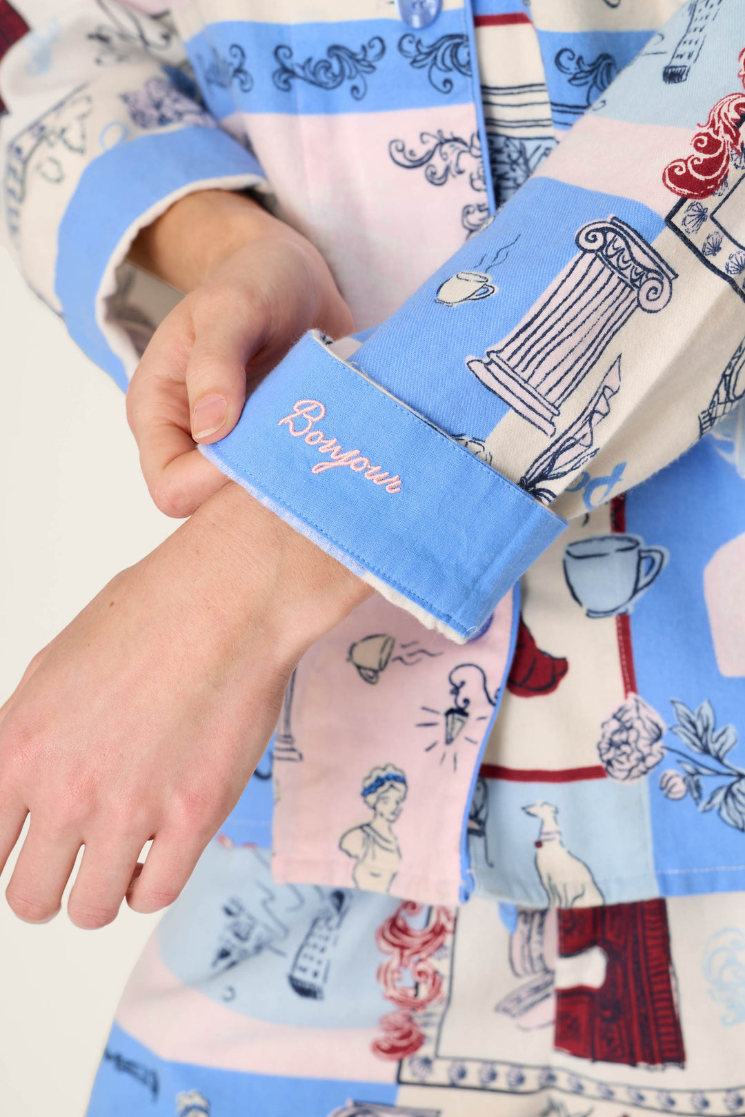 A person adjusts the cuff of their sleeve from the P.J. Salvage Allons-y Parisian Print 3-Piece Flannel PJ Set, which features a blue pattern with "Bonjour" embroidered in white script and pastel designs of columns, statues, and teacups on the flannel fabric.