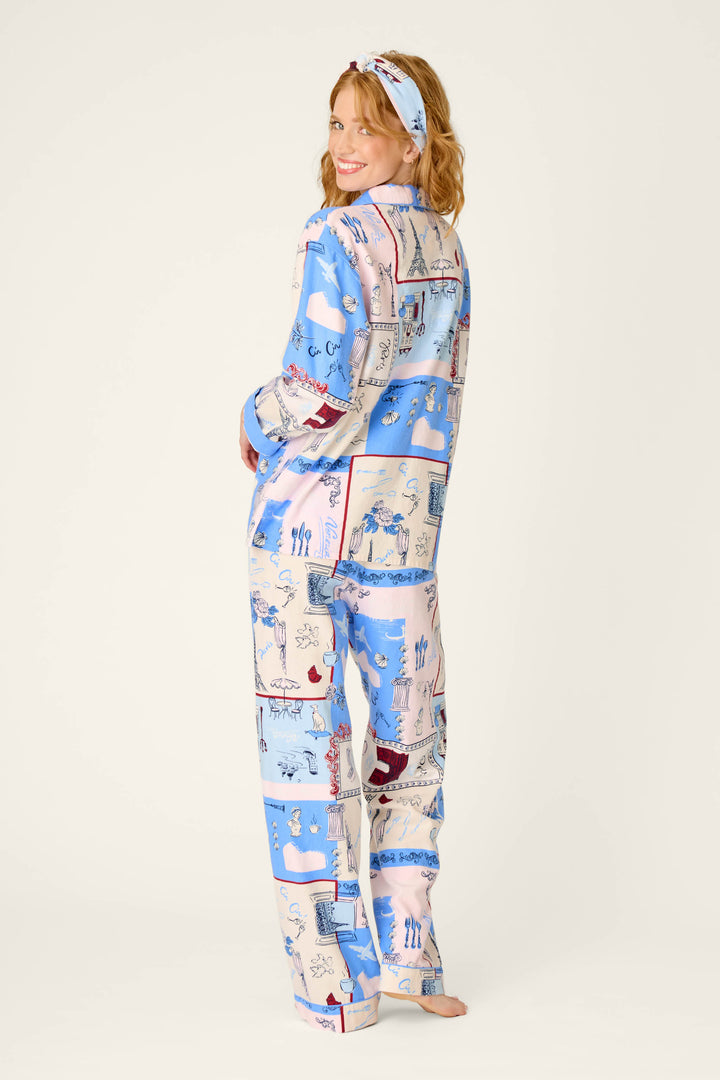 A woman smiles over her shoulder, wearing the P.J. Salvage Allons-y Parisian Print 3-Piece Flannel PJ Set, which features colorful illustrated patterns in pastel blue, pink, and beige across the pajama pants, matching long-sleeve top, and headband.