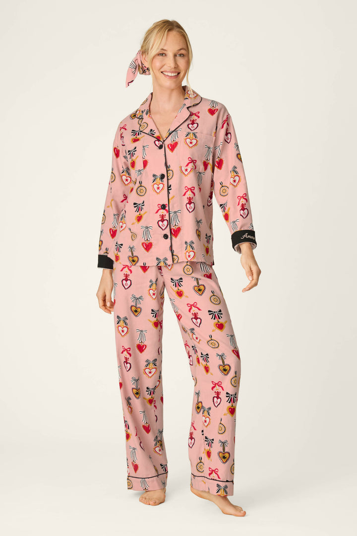 A woman smiles and stands barefoot against a plain white background, wearing the P.J. Salvage Love Lockets Print 3-Piece Long Sleeve Flannel PJ Set—pink pajamas with a colorful pattern, black trim, and a matching headscarf.