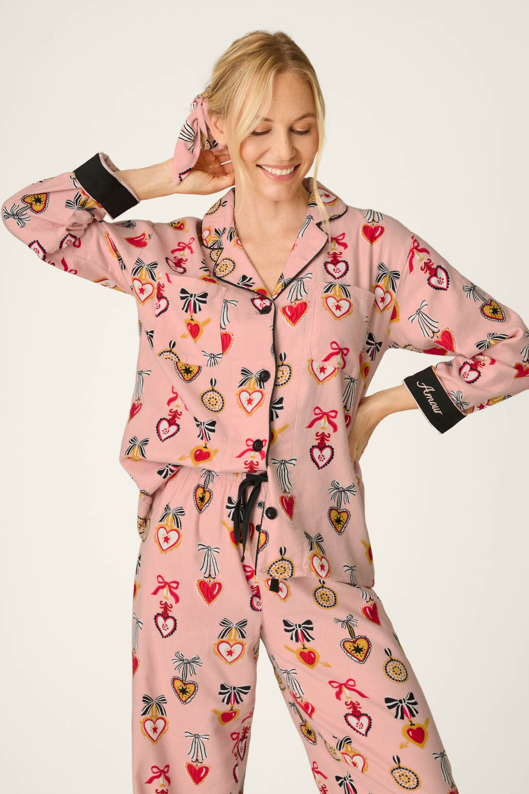A woman smiles with her eyes closed, holding her hair with one hand while wearing the P.J. Salvage Love Lockets Print 3-Piece Long Sleeve Flannel PJ Set in pink, featuring colorful heart and medal patterns. The pajama sleeve has black cuffs with white text. She stands against a plain, light-colored background.