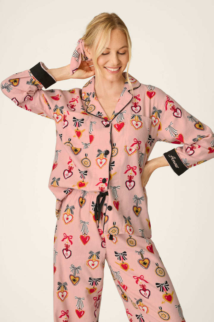 A woman smiles with her eyes closed, holding her hair with one hand while wearing the P.J. Salvage Love Lockets Print 3-Piece Long Sleeve Flannel PJ Set in pink, featuring colorful heart and medal patterns. The pajama sleeve has black cuffs with white text. She stands against a plain, light-colored background.