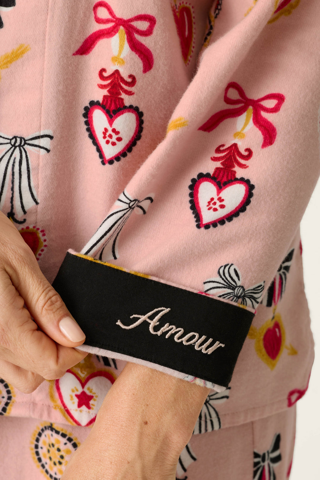 A person wears the P.J. Salvage Love Lockets Print 3-Piece Long Sleeve Flannel PJ Set, featuring pink pajamas adorned with heart and bow patterns, and holds a sleeve finished with a black cuff embroidered with “Amour.”.