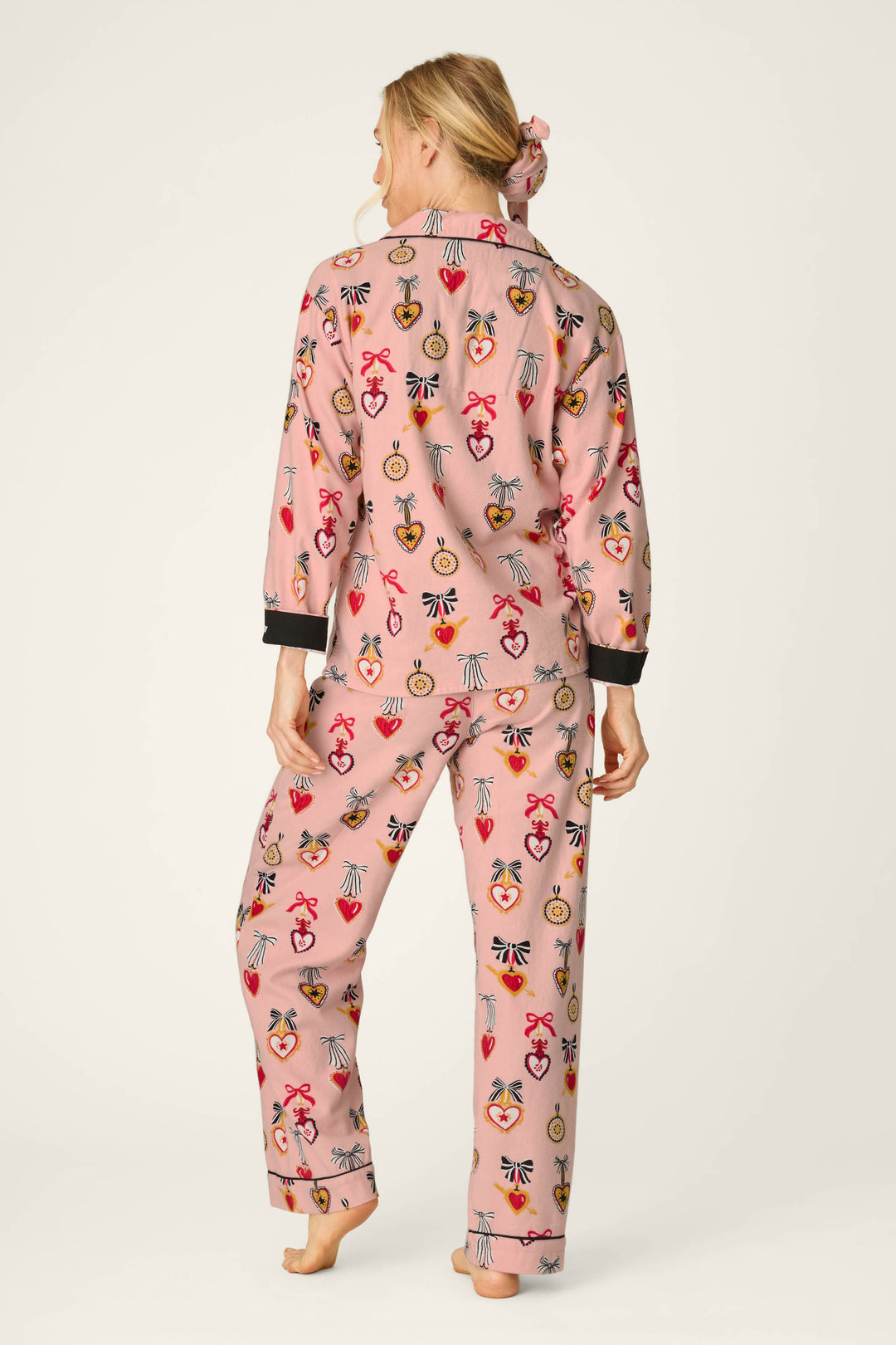 A woman stands barefoot, facing away, wearing the P.J. Salvage Love Lockets Print 3-Piece Long Sleeve Flannel PJ Set featuring colorful, playful heart and key designs. Her hair is tied back with a matching scrunchie from the set. The background is plain and light.