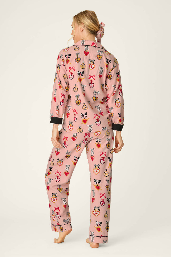 A woman stands barefoot, facing away, wearing the P.J. Salvage Love Lockets Print 3-Piece Long Sleeve Flannel PJ Set featuring colorful, playful heart and key designs. Her hair is tied back with a matching scrunchie from the set. The background is plain and light.
