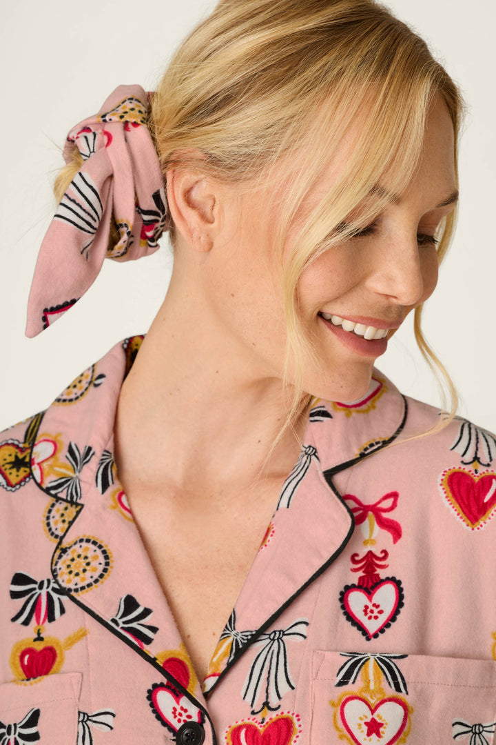 A woman with blonde hair styled in a loose side bun secured with a matching pink scrunchie is smiling and looking down, wearing the P.J. Salvage Love Lockets Print 3-Piece Long Sleeve Flannel PJ Set—a cozy pink ensemble adorned with colorful heart and bow patterns.