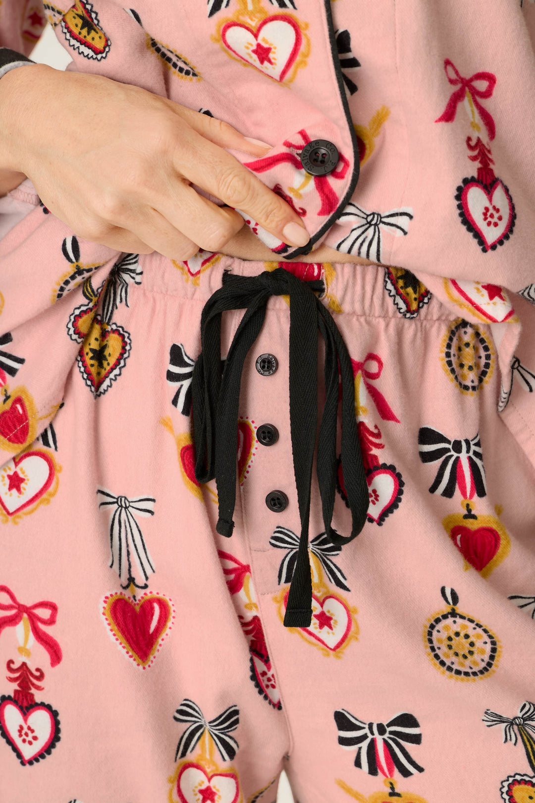 Wearing the P.J. Salvage Love Lockets Print 3-Piece Long Sleeve Flannel PJ Set, someone adjusts the button on the pink pajama top adorned with red heart and bow patterns, paired with matching drawstring pants featuring black buttons and black strings.