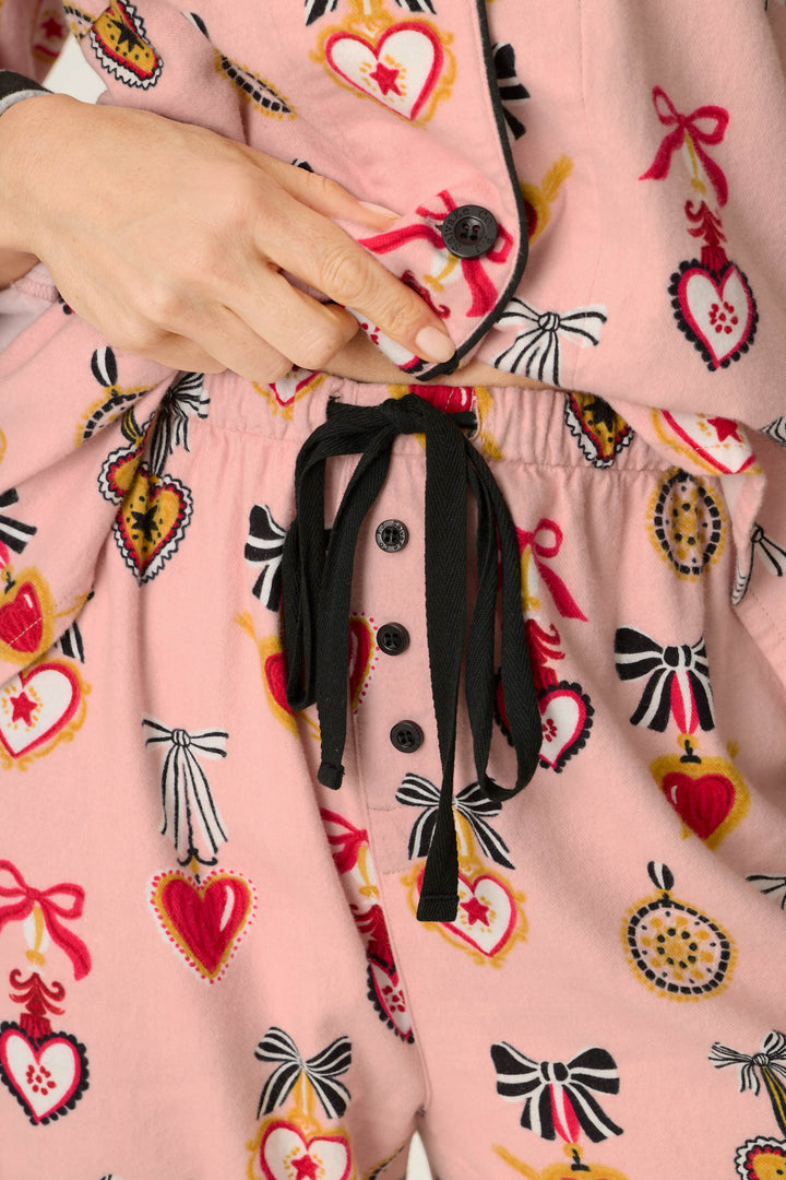 Wearing the P.J. Salvage Love Lockets Print 3-Piece Long Sleeve Flannel PJ Set, someone adjusts the button on the pink pajama top adorned with red heart and bow patterns, paired with matching drawstring pants featuring black buttons and black strings.