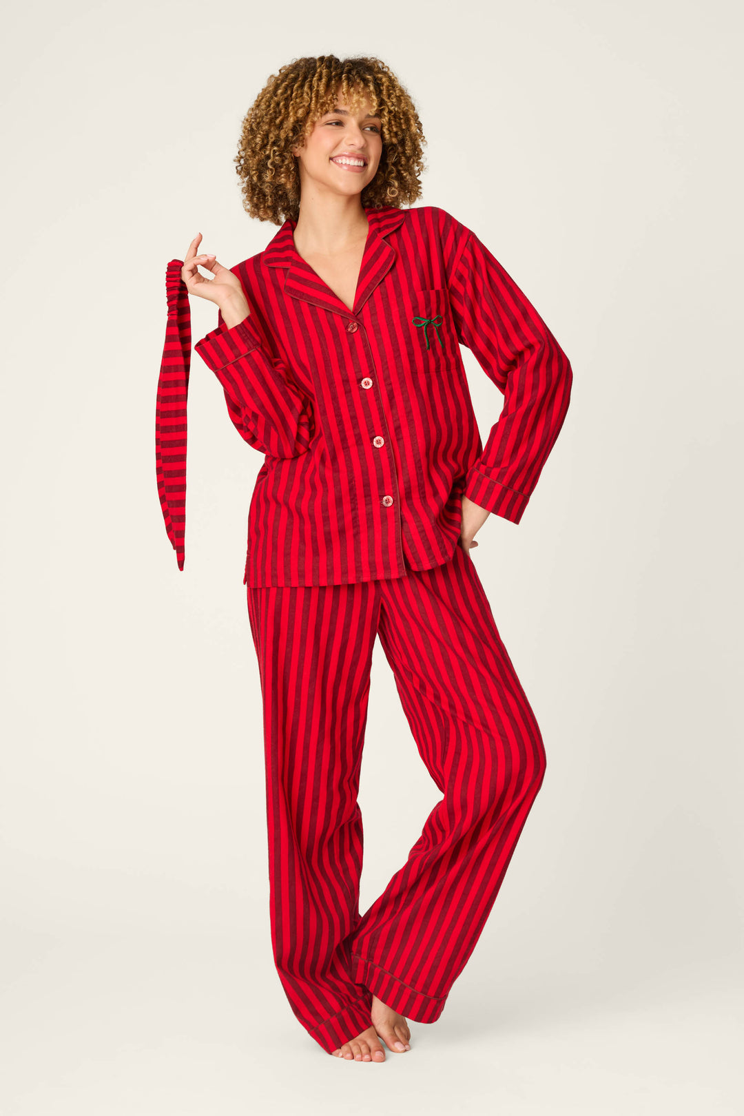 A person smiles while wearing the P.J. Salvage Bow Icon 3-Piece Long Sleeve Flannel PJ Set, featuring matching red and black stripes, and holds a striped fabric belt in one hand, standing barefoot against a plain light background.