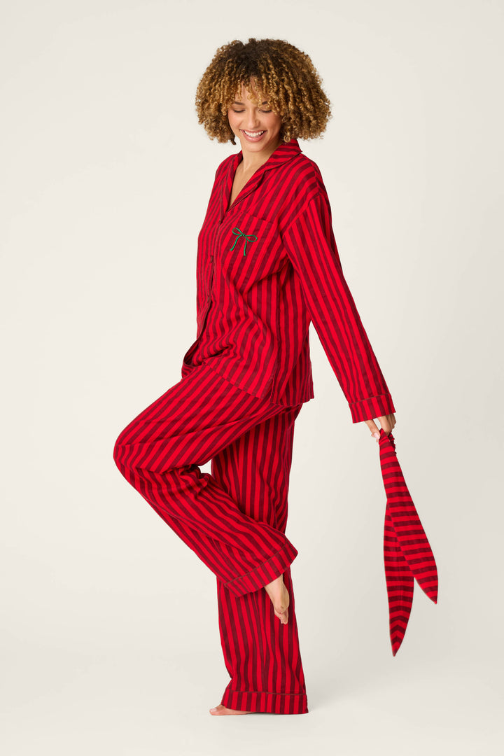 A person with curly hair wears the P.J. Salvage Bow Icon 3-Piece Long Sleeve Flannel PJ Set, smiling and lifting one foot while holding the matching striped tie against a plain background.