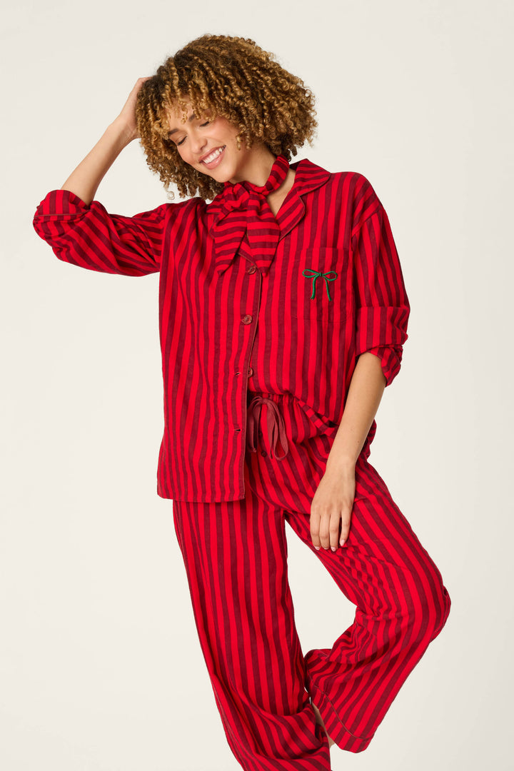 Smiling woman wearing the P.J. Salvage Bow Icon 3-Piece Long Sleeve Flannel PJ Set in red and black stripes, featuring a large matching neck bow, standing with one hand on her head and the other resting on her leg.