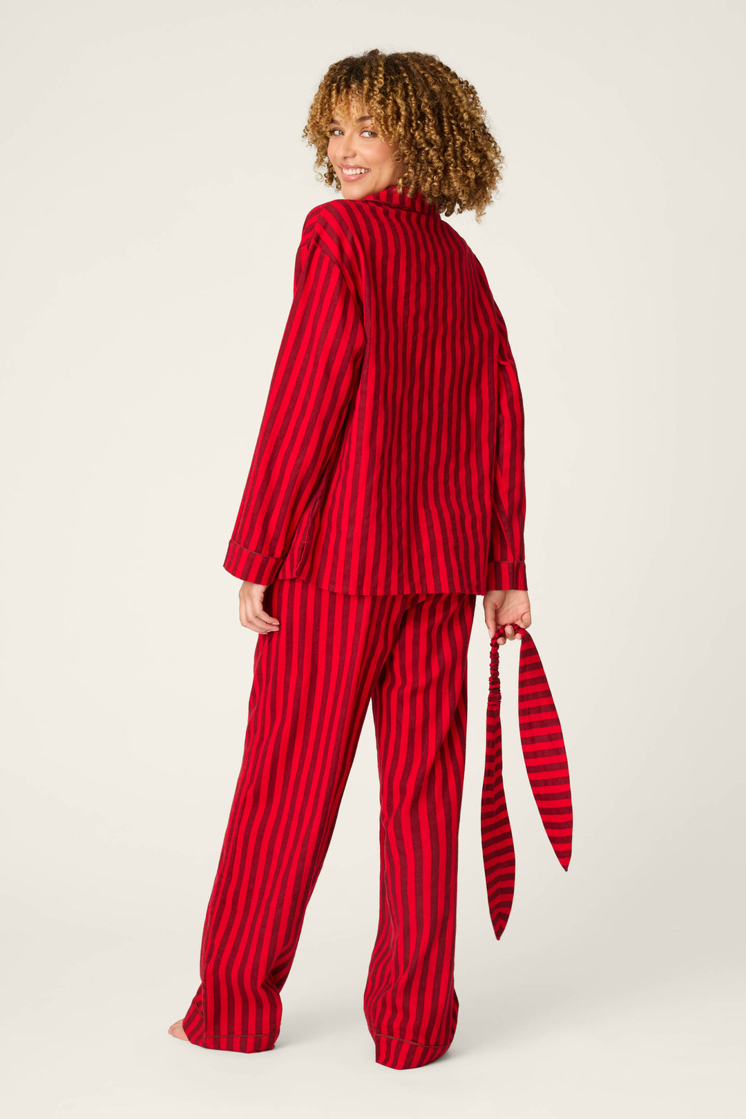 A person with curly hair, barefoot and facing away against a plain light background, is wearing the P.J. Salvage Bow Icon 3-Piece Long Sleeve Flannel PJ Set—loose red pajamas with vertical black stripes—and holding the matching striped belt in one hand.
