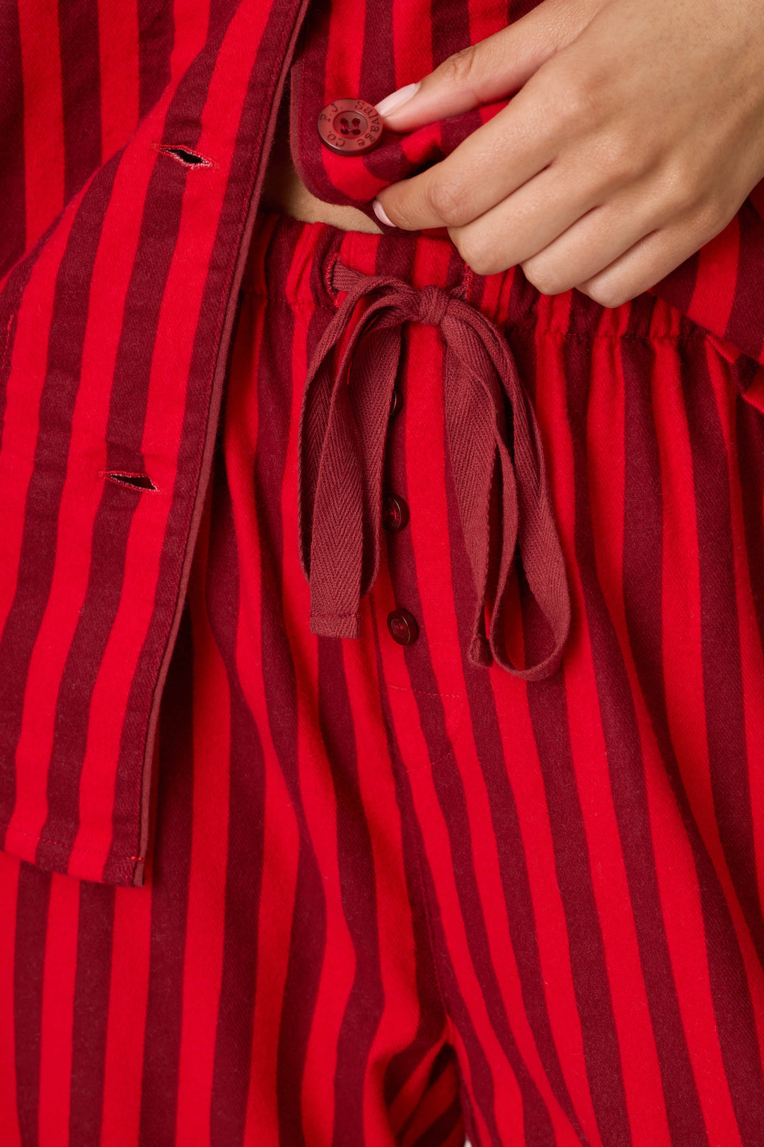 A close-up view shows a person’s hand unbuttoning the red and dark red striped top from the P.J. Salvage Bow Icon 3-Piece Long Sleeve Flannel PJ Set, with matching drawstring striped pants featuring button details.