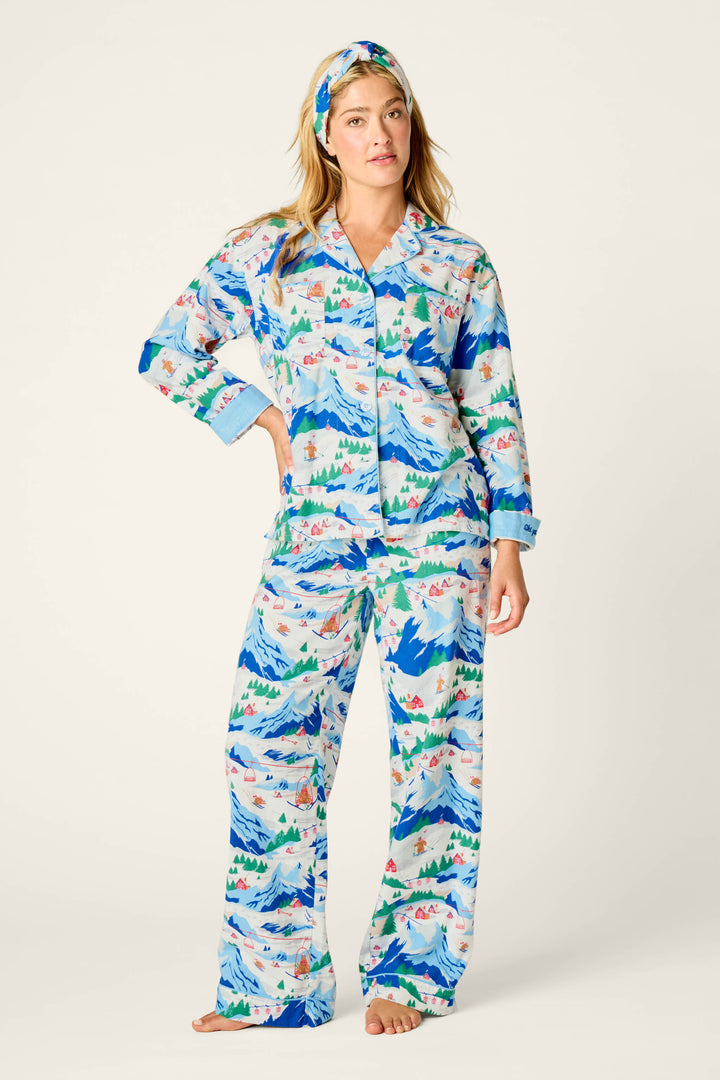 A woman stands barefoot wearing the P.J. Salvage Ski Ya Later Skiing Bears Print 3-Piece Flannel PJ Pant Set, which features matching pajamas and a headband adorned with a colorful nature-inspired print of skiing bears, mountains, trees, and tents on a white background.