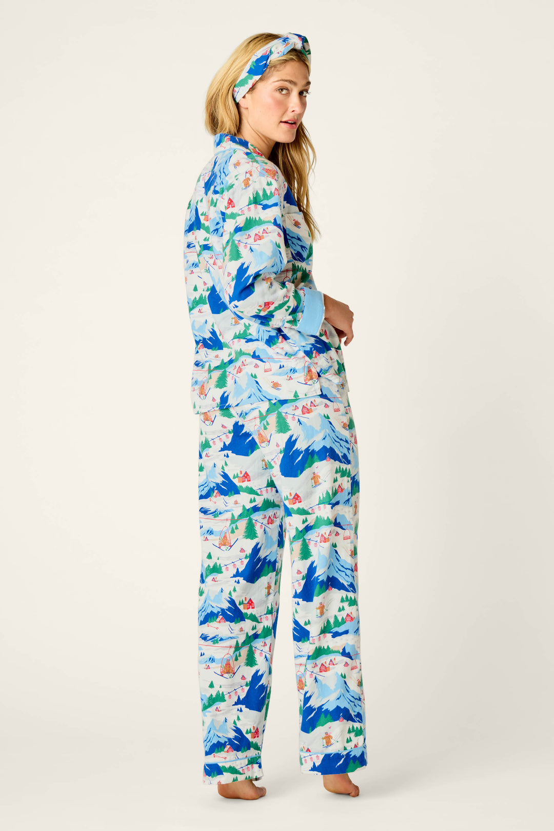 A woman wearing the P.J. Salvage Ski Ya Later Skiing Bears Print 3-Piece Flannel PJ Pant Set, including matching pajamas and headband, stands barefoot with her body angled slightly away from the camera against a simple, light backdrop.